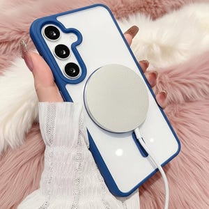 以下が含まれることがあります： 白と青の携帯電話ケースに、銀色の円形磁気充電器が取り付けられています。ケースは青い縁と透明な背面を備えています。充電器は白いケーブルに接続されています。ケースはピンクと白のふわふわした表面に置かれています。