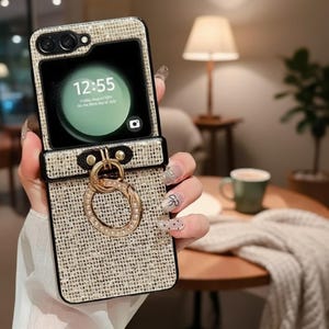 May include: A beige and black phone case with a gold ring holder. The case is open, revealing a phone screen displaying the time and date. The case has a textured, woven appearance and a black leather strap.