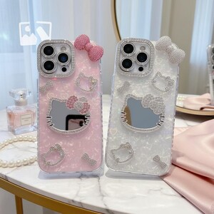 May include: Two phone cases, one pink and one white, adorned with sparkling embellishments. Each case features a Hello Kitty design with a mirrored face, a bow, and additional decorative elements. The cases are designed to protect the phone's camera.