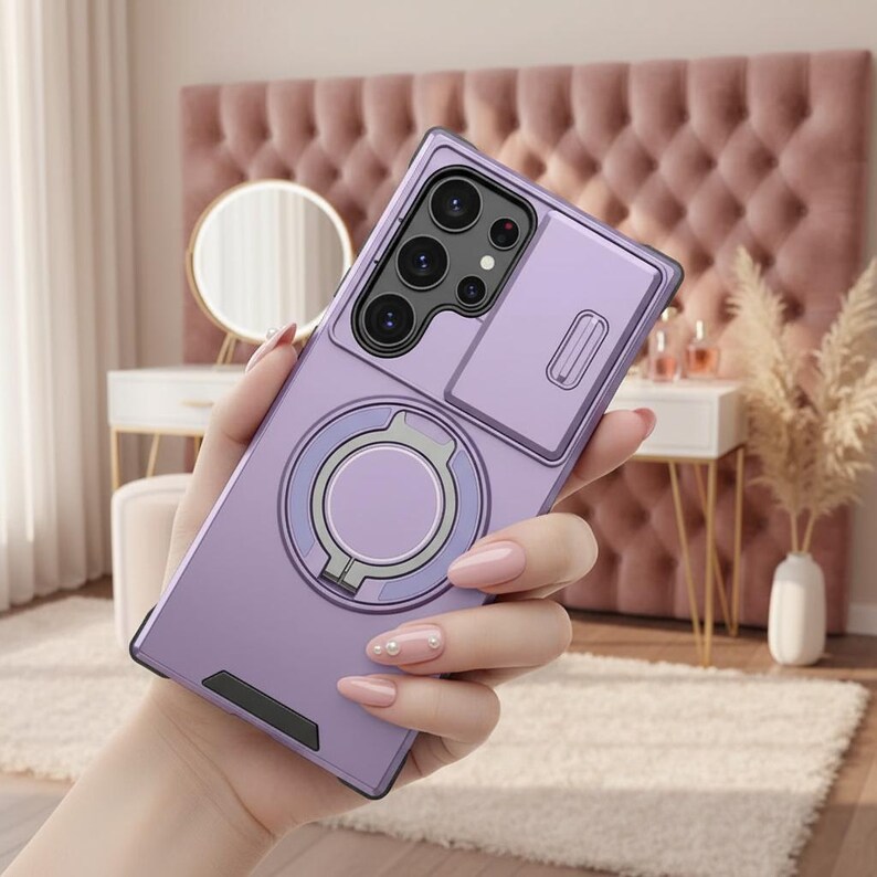 May include: A lavender phone case with a camera protector and a ring holder. The case has a sleek design and a built-in stand. The ring holder is silver and gray. The phone case is held in a hand with pink nail polish.