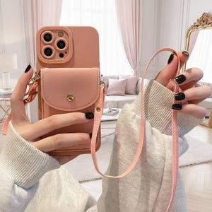 May include: A blush pink phone case with a small, matching wallet attached. The case has a strap for carrying. The phone case has a camera lens protector and a gold-colored button closure on the wallet.