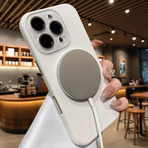 Puede incluir: Funda de teléfono blanca con un accesorio magnético circular gris. La funda tiene un protector de lente de cámara transparente. Un cable de carga blanco está conectado al accesorio magnético. La funda del teléfono está sobre un soporte blanco.