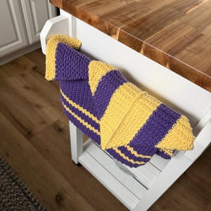May include: A rectangular crochet kitchen towel featuring alternating vertical stripes of deep purple and bright yellow yarn. The textile has a textured, woven appearance with a consistent stitch pattern throughout, draped over the side of a white furniture piece.
