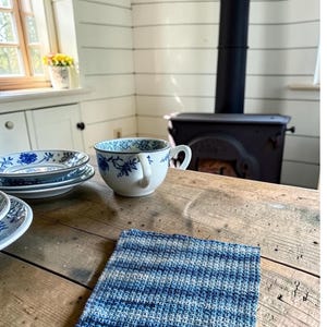 May include: A square, hand-crocheted dishcloth featuring a textured, variegated pattern of horizontal stripes in various shades of blue, ranging from light sky blue to deep navy, with a consistent, dense stitch throughout the entire piece.