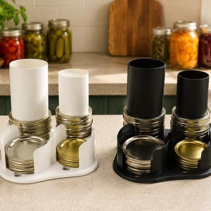 May include: Two plastic organizers for canning jar lids and rings, one white and one black. Each unit features two vertical cylindrical towers for holding rings and two open-front compartments for stacking metal lids, keeping kitchen supplies organized.