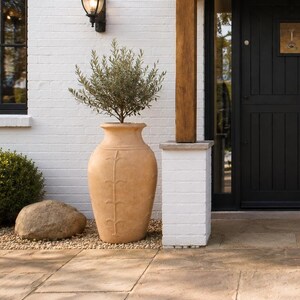 May include: A black front door with vertical panels and a brass house number plaque, framed by a wooden porch structure. Two large, light brown terracotta urns with embossed leaf patterns flank the entrance, each containing a small olive tree.