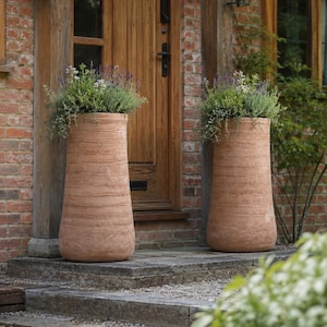 May include: Two tall, cylindrical terracotta planters with a textured, ribbed surface finish. Each planter is filled with green foliage and purple lavender flowers, positioned on either side of a wooden door set in a red brick wall.