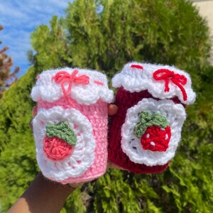Pode incluir: Duas capas de crochê para canecas, uma rosa e outra vermelha, que se assemelham a potes de geleia. Cada capa apresenta uma borda branca com vieiras e uma aplicação branca com um morango de crochê. A capa rosa tem um laço rosa, e a vermelha tem um laço vermelho.