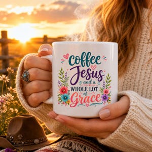 May include: A white ceramic mug featuring the text "coffee Jesus and a WHOLE LOT of Grace" in colorful script and block lettering, surrounded by a decorative floral design with small pink hearts and green leaves on the front surface.