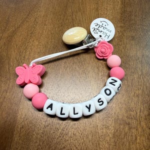 以下が含まれることがあります： 白い文字ビーズで「ALLYSON」と綴られたピンクと白のおしゃぶりクリップ。クリップにはピンクの蝶、ピンクのバラ、ピンクのビーズが付いています。「Hand made」と書かれたタグが添付されています。