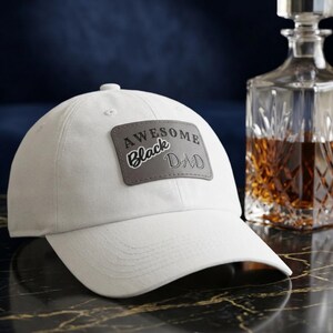 Peut inclure: Casquette de baseball blanche avec une visière incurvée. Le chapeau comporte un patch gris avec les mots "AWESOME Black DAD" en lettres noires. Un décanteur en verre avec du liquide ambré est en arrière-plan.