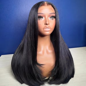 Può includere: Parrucca nera a capelli lisci acconciata su una testa di manichino. La parrucca presenta un'attaccatura dei capelli dall'aspetto naturale e ciocche lunghe e lisce. Il manichino ha il trucco applicato, tra cui ombretto e rossetto. Lo sfondo è blu.