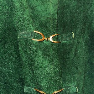 May include: Close-up of a textured, emerald green fabric with three sets of decorative gold-colored clasps. The clasps are curved and attached to the fabric with small, rectangular straps. The fabric has a slightly worn appearance.