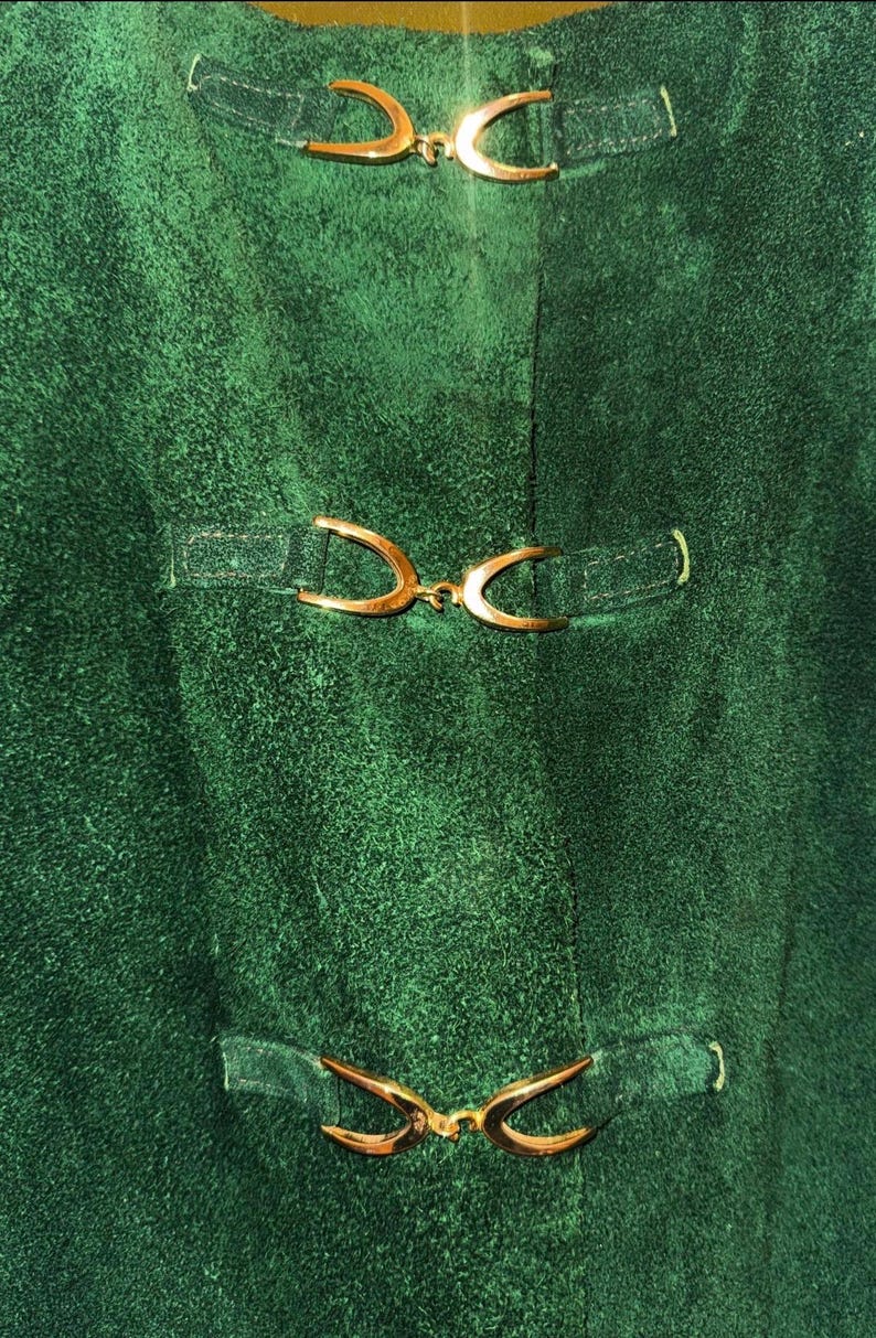 May include: Close-up of a textured, emerald green fabric with three sets of decorative gold-colored clasps. The clasps are curved and attached to the fabric with small, rectangular straps. The fabric has a slightly worn appearance.