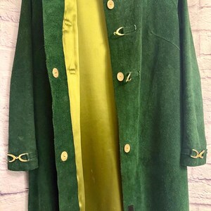 May include: A long, dark green suede coat with a light green satin lining. The coat features gold-colored clasp closures and decorative accents on the sleeves. The coat is open, revealing the interior lining.