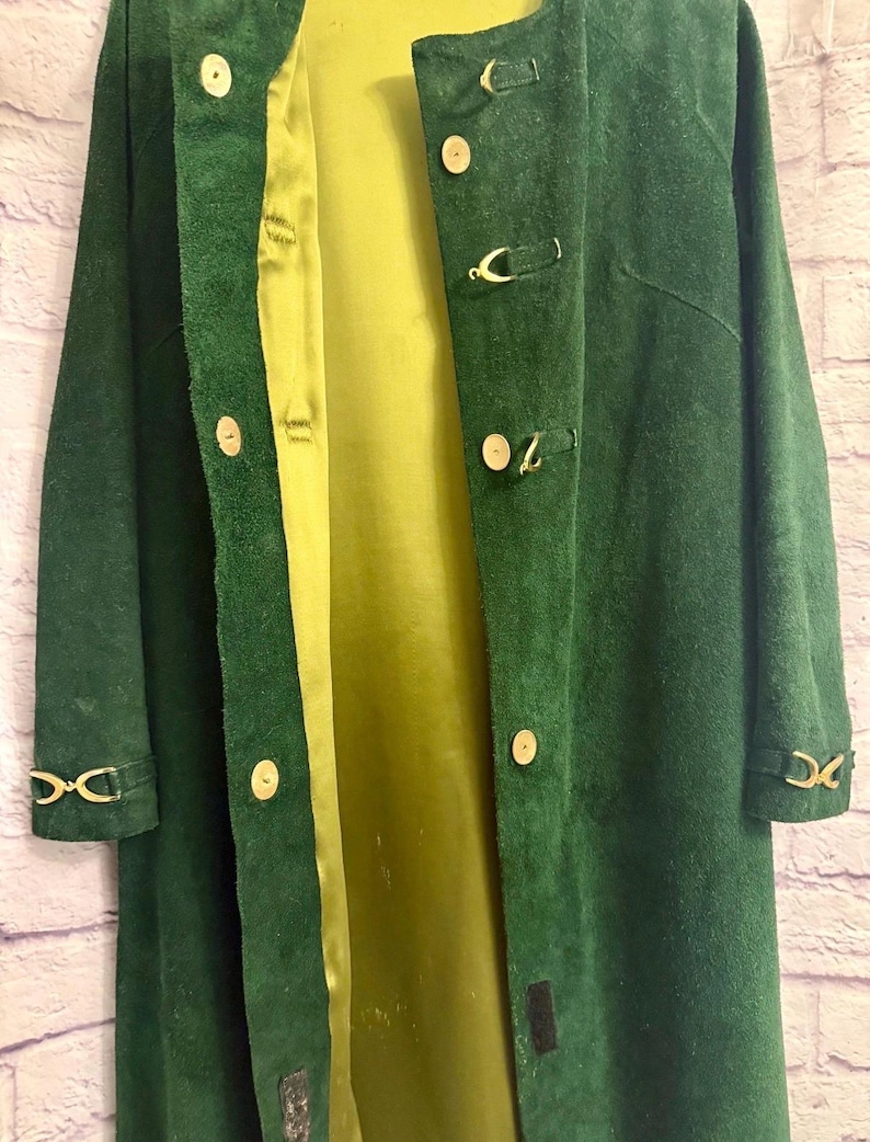 May include: A long, dark green suede coat with a light green satin lining. The coat features gold-colored clasp closures and decorative accents on the sleeves. The coat is open, revealing the interior lining.