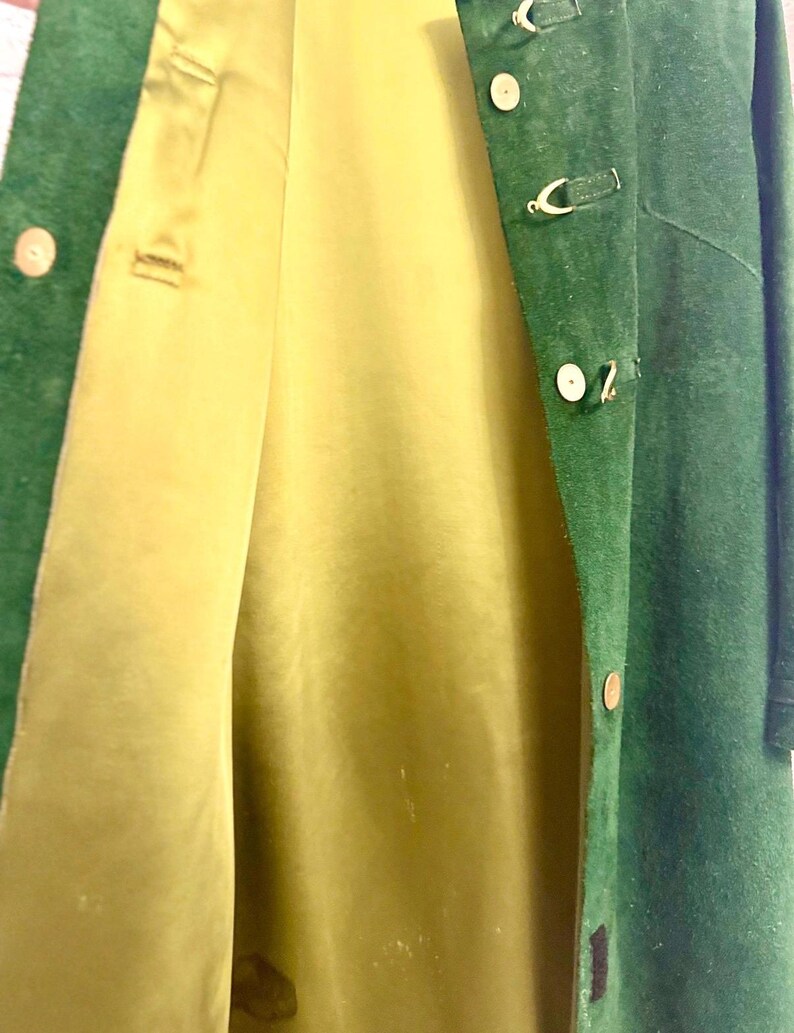 May include: A green suede coat with a light yellow lining. The coat has a button closure and decorative gold-colored clasps. The coat is open, revealing the interior lining.