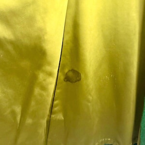May include: Close-up of a lime green satin fabric with a darker stain. The fabric is folded, revealing a glimpse of a green trim. The texture of the satin is visible, with subtle variations in color and sheen.