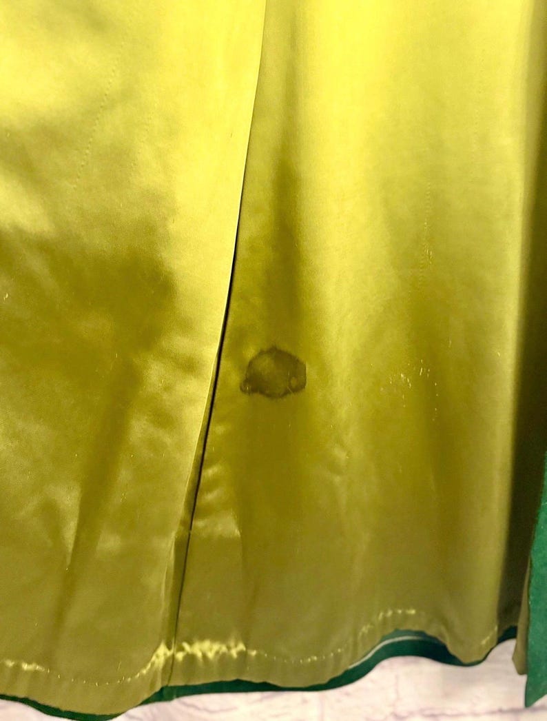 May include: Close-up of a lime green satin fabric with a darker stain. The fabric is folded, revealing a glimpse of a green trim. The texture of the satin is visible, with subtle variations in color and sheen.