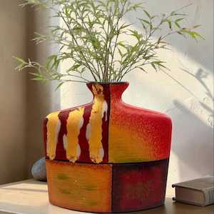 Puede incluir: Un jarrón de cerámica ancho y plano con una paleta de colores vibrantes en rojo, naranja y amarillo. La superficie muestra bloques texturizados pintados a mano y pinceladas verticales en tono dorado. Una planta verde está dentro del jarrón.