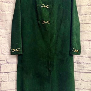 May include: A long, dark green suede coat with gold-colored metal clasps. The coat has long sleeves and a simple, elegant design. The interior lining is a light yellow color. A vintage outerwear piece.