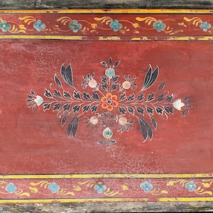 Può includere: Una scatola rettangolare in legno rosso antico con un design floreale. La scatola presenta un motivo floreale centrale con foglie nere, fiori arancioni e bianchi e un bordo con fiori blu e dettagli gialli. La scatola viene probabilmente utilizzata per la conservazione.
