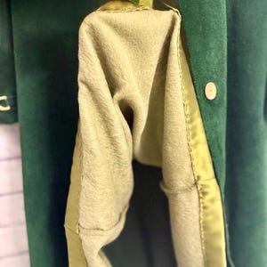 May include: Close-up of a dark green coat with a light olive-green lining. The coat has a button closure and a satin trim along the edges. The interior lining is a soft, fuzzy material.