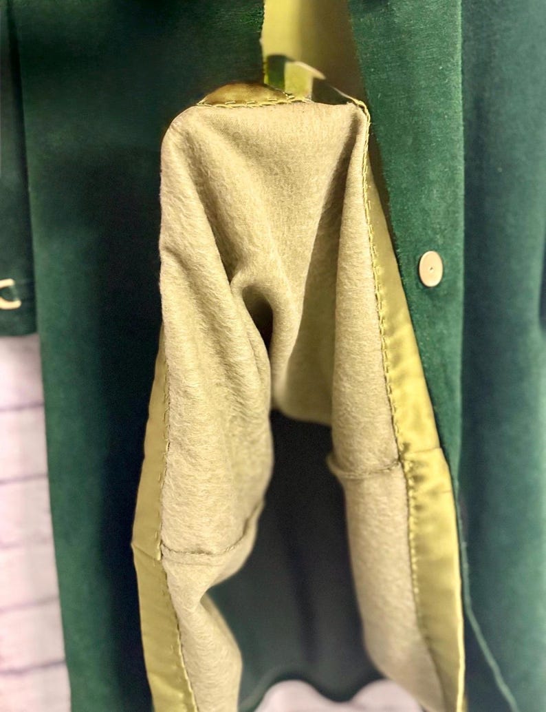 May include: Close-up of a dark green coat with a light olive-green lining. The coat has a button closure and a satin trim along the edges. The interior lining is a soft, fuzzy material.