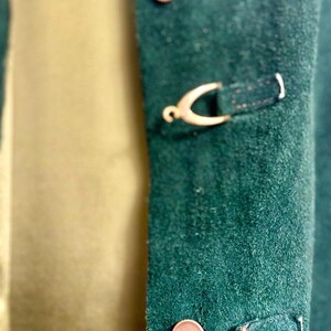 May include: Close-up of a dark green suede jacket with gold-colored metal clasps and buttons. The jacket has a classic design with a soft, textured appearance. The buttons and clasps add a touch of elegance to the garment.