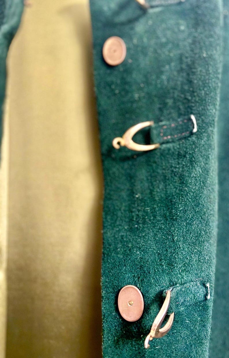 May include: Close-up of a dark green suede jacket with gold-colored metal clasps and buttons. The jacket has a classic design with a soft, textured appearance. The buttons and clasps add a touch of elegance to the garment.