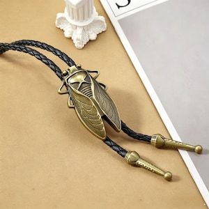 May include: A bolo tie with a bronze-toned cicada pendant, showcasing intricate wing details. The pendant is suspended from a black, braided leather cord, finished with decorative tips. The tie is displayed on a neutral surface.