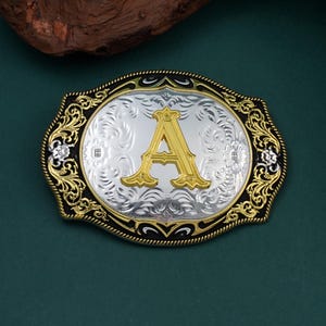 May include: An oval-shaped silver and gold belt buckle with the letter "A" in gold. The buckle has an ornate floral design around the edges and a black border. The background is a dark green.