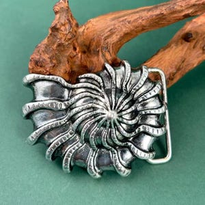 May include: A silver-coloured belt buckle featuring a spiral design. The buckle has a rectangular frame and a detailed, textured pattern resembling a nautilus shell. The background is a muted green.