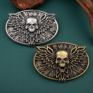 May include: Two oval-shaped belt buckles with skull and wing designs. One is silver-toned, the other is bronze-toned. The skulls have open mouths and are centered on the buckles, with detailed wing patterns surrounding them. The buckles are on a dark green surface.