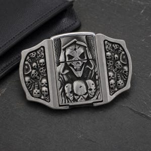 Könnte beinhalten: Ein silberfarbenes Feuerzeuggehäuse aus Metall mit einem detaillierten Reliefdesign einer zentralen Figur mit Kapuzenschädel, umgeben von zahlreichen kleineren Schädeln auf den Seitenteilen. Die Oberfläche hat ein verwittertes, antikes Finish.