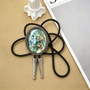 May include: Oval bolo tie with a shimmering abalone shell center, framed by a silver-toned border. The black braided cord is connected to decorative silver-toned tips. A unique accessory.