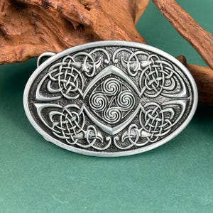 May include: Oval silver belt buckle with Celtic knot designs. The buckle features intricate knotwork patterns and a central triskele symbol. The metal buckle has a textured surface and a classic, vintage aesthetic.