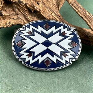 May include: Oval belt buckle with a geometric pattern. The buckle has a navy blue base with white and maroon details. It is framed by a silver-coloured metal border. The buckle is likely made of metal and is suitable for a belt.