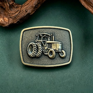 Puede incluir: Hebilla de cinturón rectangular de color latón antiguo con un diseño detallado de tractor. El tractor tiene neumáticos traseros grandes y una cabina, sobre un fondo texturizado. Un accesorio clásico.