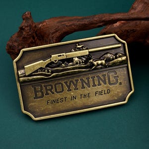 May include: A rectangular, antique brass-colored belt buckle featuring a rifle, deer, and mountain scene. The word "BROWNING" is prominently displayed, with "FINEST IN THE FIELD" below. The buckle has a detailed, embossed design.