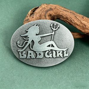 May include: Oval-shaped silver-toned metal belt buckle featuring a devilish woman with a trident and the words "BAD GIRL". The buckle has a textured background and is set against a natural wood element.