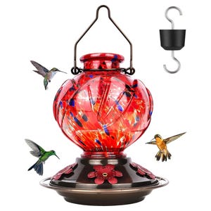 May include: A red glass hummingbird feeder with a decorative design, a dark metal base, and a hanging hook. The feeder is surrounded by three hummingbirds in flight. Includes a black hook.