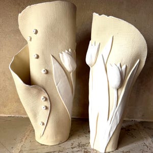 Könnte beinhalten: Zwei cremefarbene Keramikvasen mit einer strukturierten, stoffähnlichen Oberfläche. Die linke Vase ist mit kleinen erhabenen runden Perlen verziert, während beide Vasen weiße, skulpturale Tulpenmotive mit Stängeln und Blättern aufweisen.