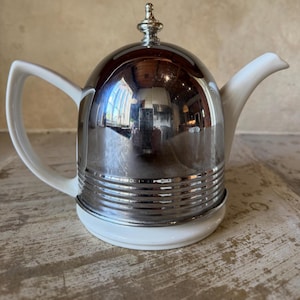 Op de afbeelding: Een vintage theepot met een glanzende, zilverkleurige metalen koepel en een witte keramische basis, handvat en tuit. De theepot heeft horizontale ribbels rond de basis en een decoratieve bekroning bovenop. Het reflecterende oppervlak weerspiegelt de omgeving.