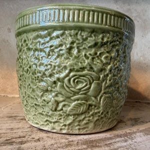Op de afbeelding: Een saliegroene keramische plantenpot met een textuurrijk, hobbelig oppervlak en een reliëf van een roos. De cilindrische pot heeft een gladde, verticaal geribbelde rand aan de bovenkant en een glanzende afwerking die het bloemmotief accentueert.