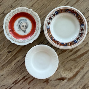 May include: Three small decorative plates. One plate features a portrait within a red border, another has a patterned rim with blue, brown, and white details, and the third is plain white.