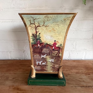 May include: A flared metal vase with a gold-toned rim and base, featuring a painted scene of two riders in red coats on horseback accompanied by several white dogs in a field with a tree, set upon a solid dark green rectangular pedestal base.