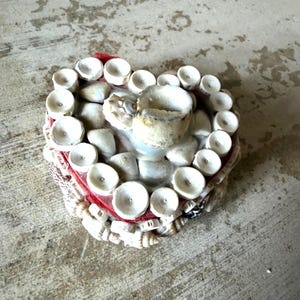 May include: Heart-shaped decorative object made of seashells, with a red border and a central cup-like structure. The object is adorned with numerous small, white seashells and sits on a textured surface.