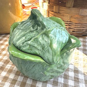 May include: A ceramic cabbage-shaped container with a lid. The container is a light green color with darker green veining details. The lid fits snugly on top of the base, creating a decorative and functional piece. The container is sitting on a checkered tablecloth.