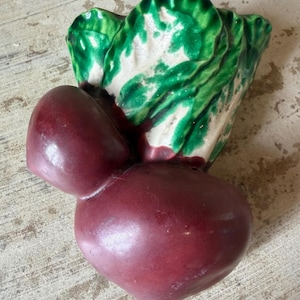 May include: A ceramic beet-shaped wall pocket with a glossy finish. The beet is a deep burgundy color, with green and white leafy tops. The decorative piece is designed to be mounted on a wall and adds a touch of whimsy to any space.